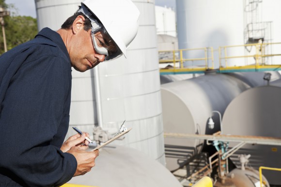 Mid adult man with clipboard at chemical plant.  Storage tanks in background.